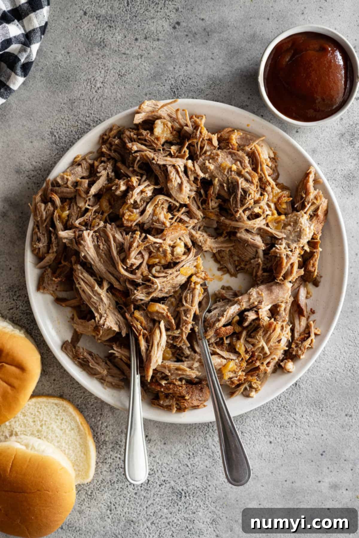 Overhead view of pulled pork on a white plate with two forks in the meat. Buns and bbq sauce off to the side. 