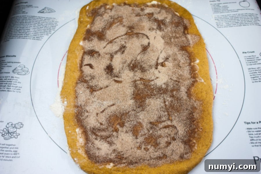 Pumpkin bread dough expertly rolled out on a pastry mat, with the sweet cinnamon-sugar filling evenly spread across its surface, ready to be rolled into a log.