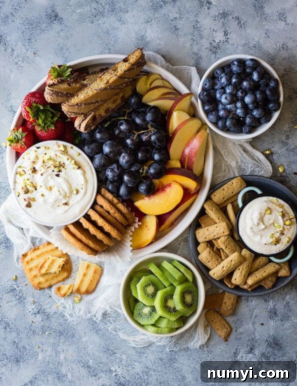A beautifully arranged fruit tray with various colorful fruits like grapes, strawberries, peaches, blueberries, and kiwi, alongside delicious dips and cookies.