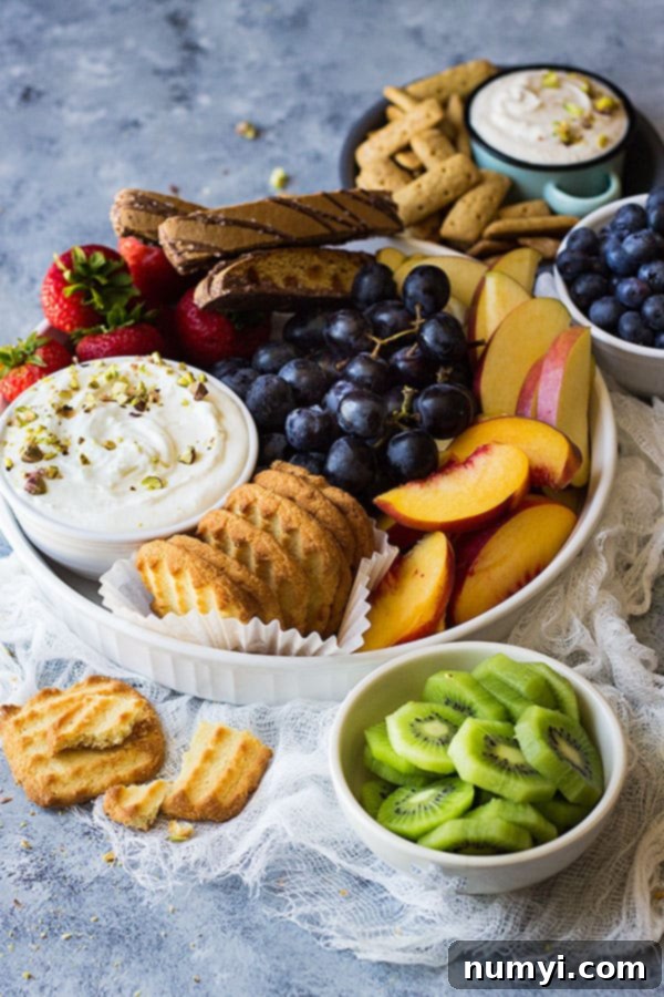 A vibrant fruit tray from an overhead perspective, displaying various colorful fruits, bowls of dips, and different types of cookies.