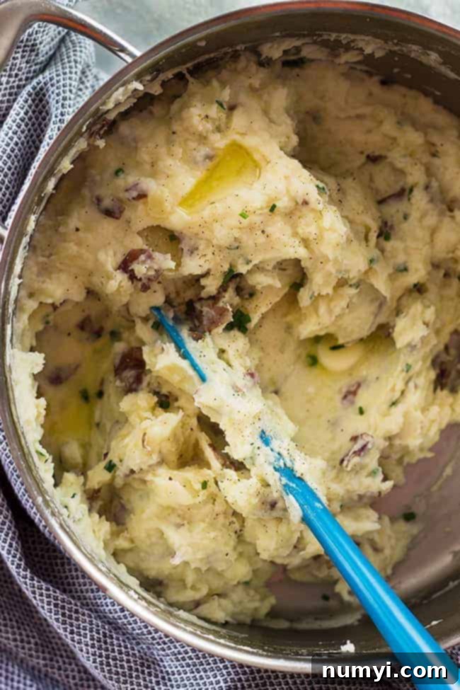Overhead closeup of a blue spatula stirring a big metal bowl of chunky mashed potatoes, highlighting their rustic texture.