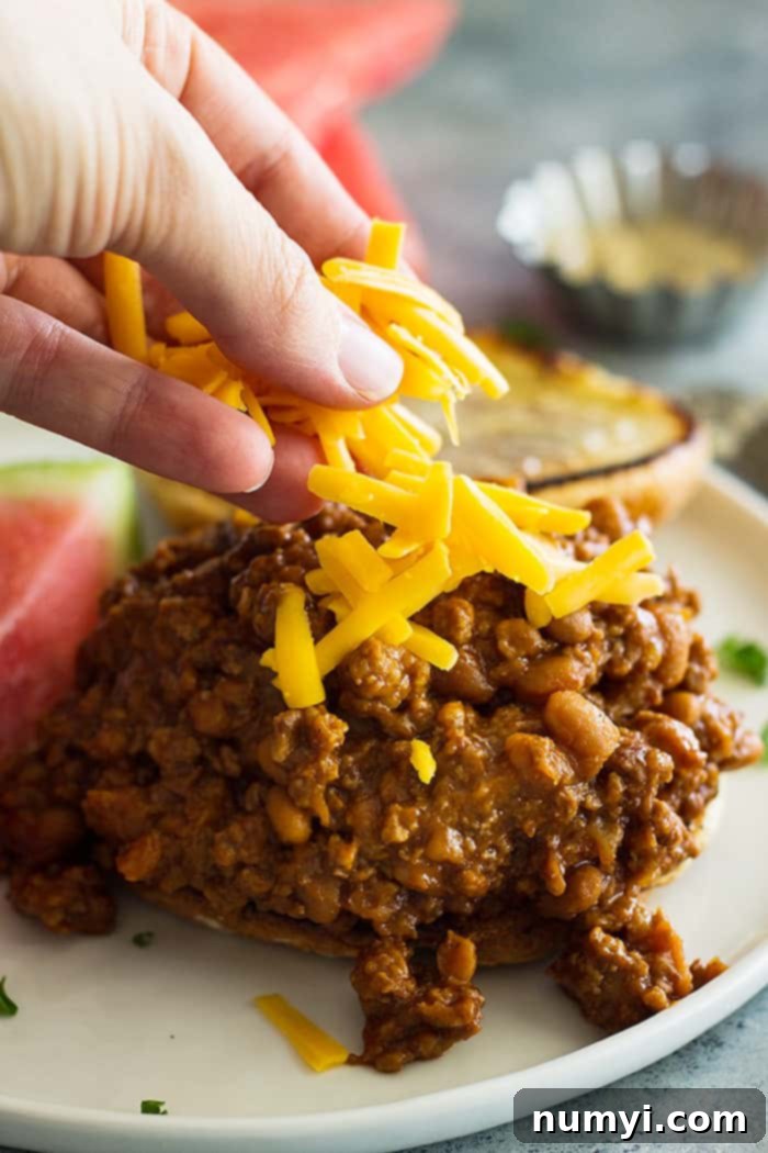 Top shot of shredded cheddar cheese being sprinkled generously on top of the slow cooker beef and bean sloppy joe mixture on a white plate.