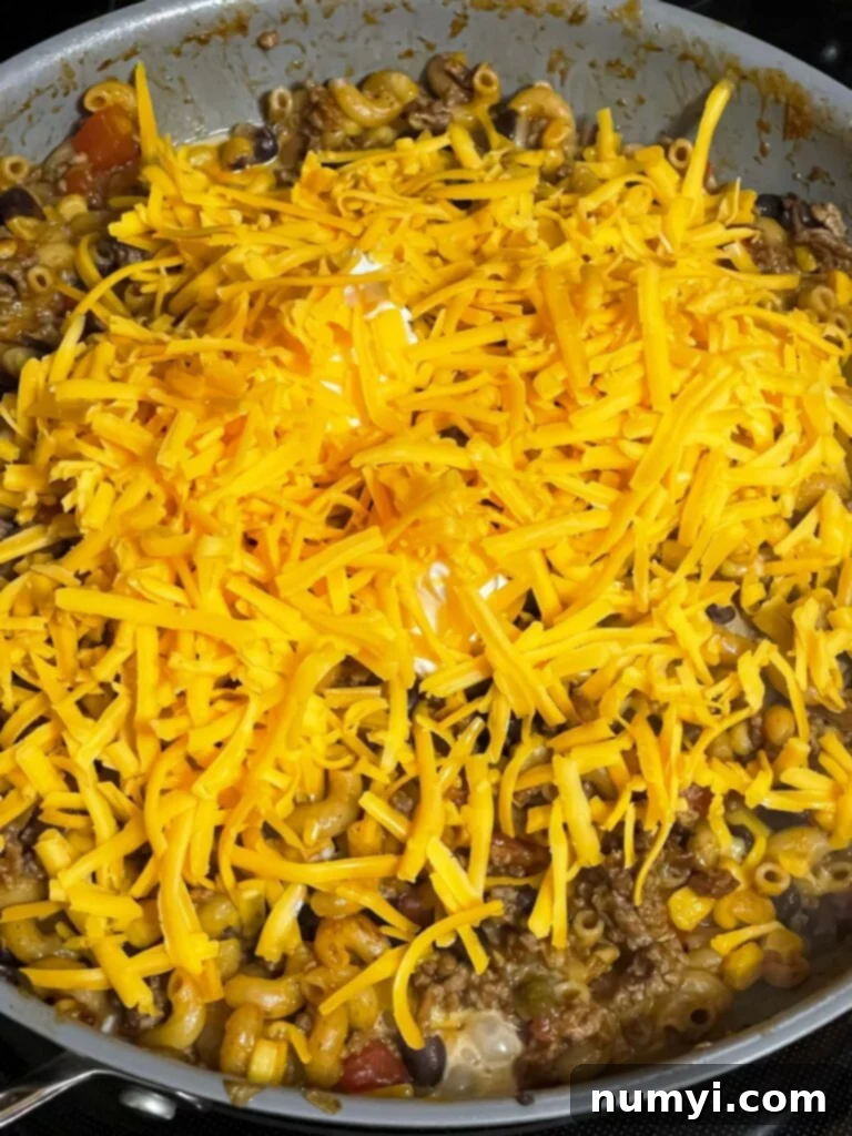 Shredded cheddar cheese being added to the cooked taco pasta mixture in a skillet.