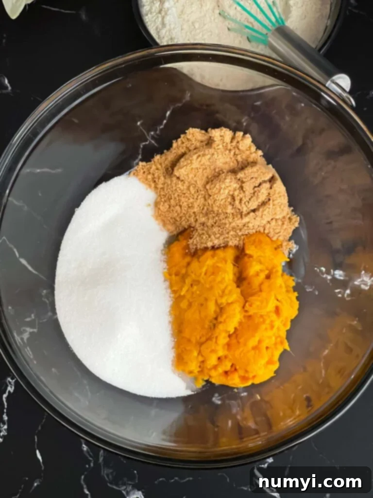 Sugars and mashed sweet potato being creamed in a glass mixing bowl on a dark surface.