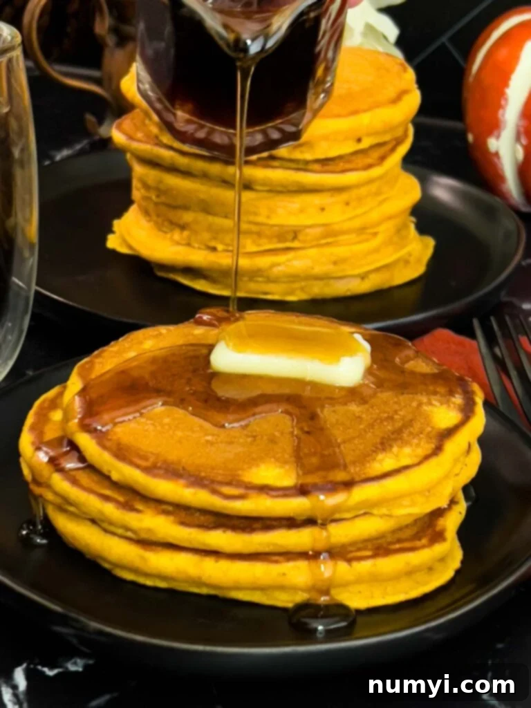 A beautifully stacked pile of fluffy pumpkin pancakes, generously topped with a pat of melting butter and a drizzle of rich maple syrup, showcasing their golden-brown edges on a rustic dark plate.