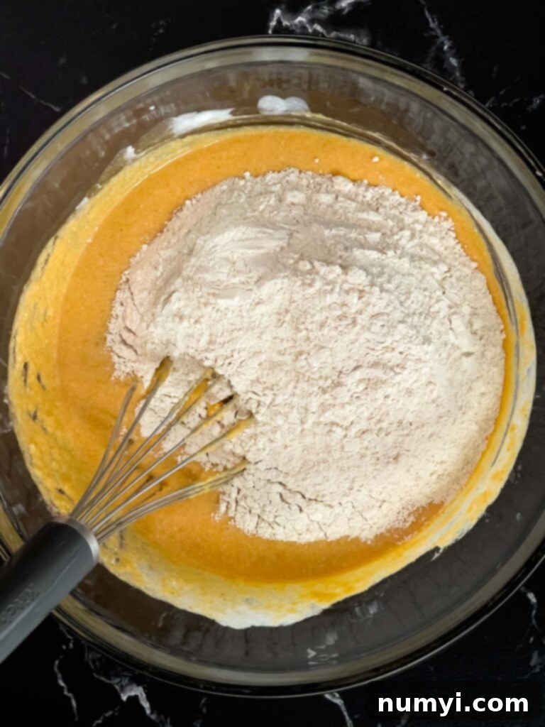 Dry ingredients being carefully added to the vibrant orange wet ingredients in a clear glass bowl, showcasing the initial mixing stage for pumpkin pancakes.