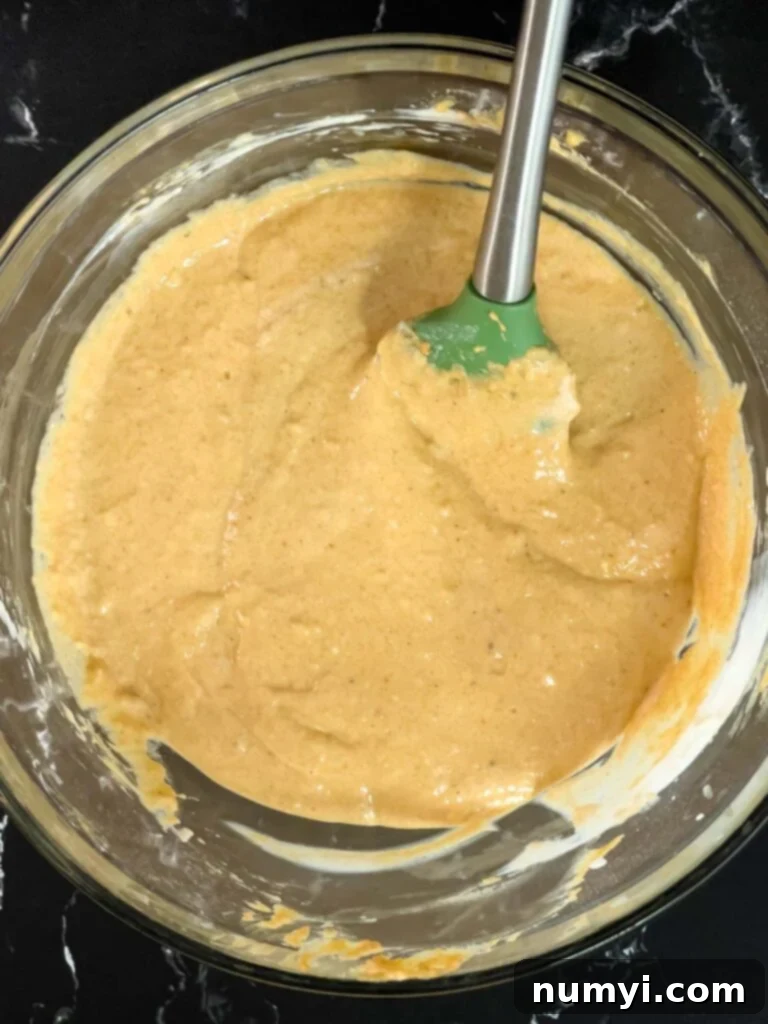 A perfectly mixed, smooth pumpkin pancake batter, rich orange in color, resting in a glass bowl on a dark textured surface, ready for cooking.