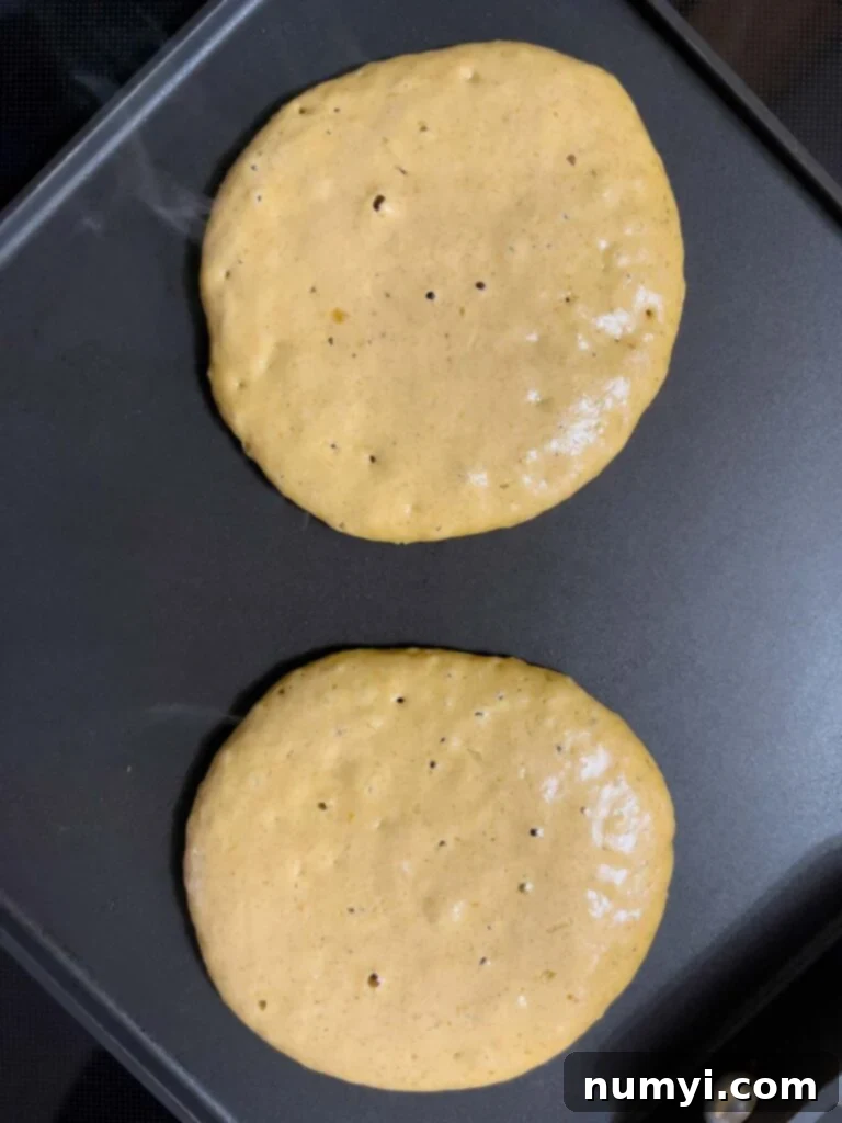 Close-up of a pumpkin pancake cooking on a griddle, showing numerous small bubbles actively popping on its surface, indicating it's nearly ready to be flipped.