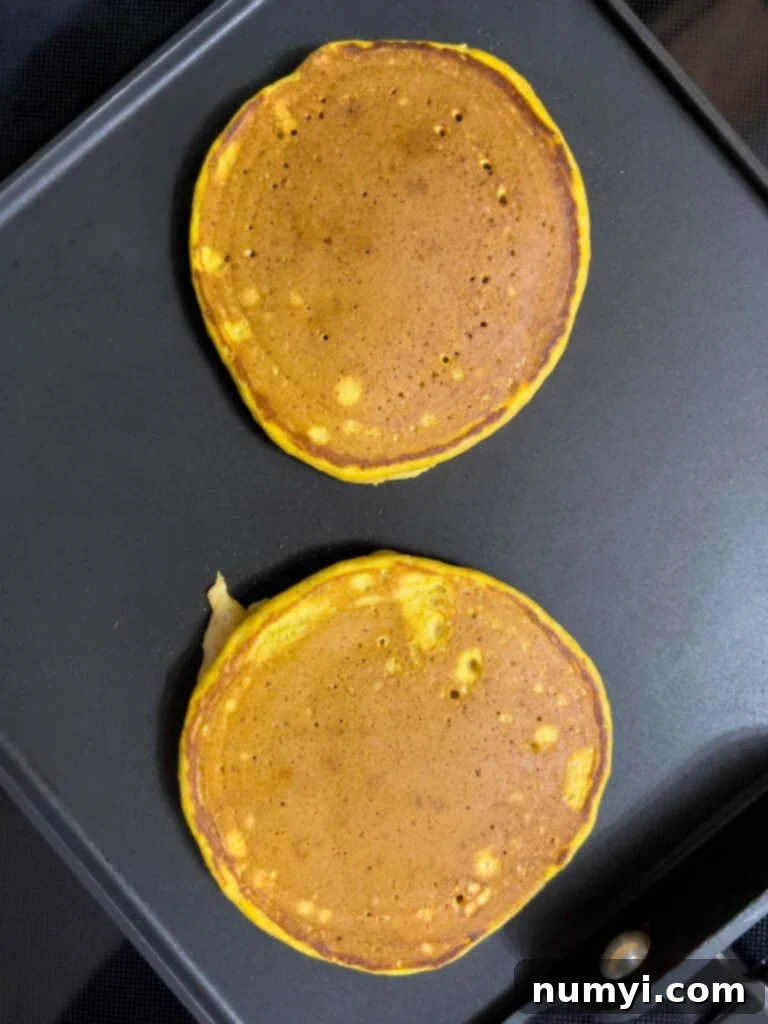 Several pumpkin pancakes cooking on a flat griddle, some with golden-brown flipped sides and others still bubbling, creating a delicious scene.