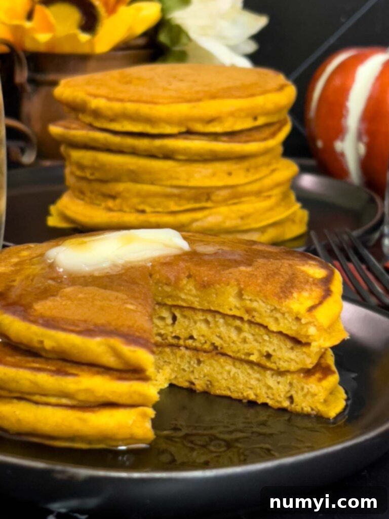 A high stack of golden-brown fluffy pumpkin pancakes, topped with melting butter and glistening with maple syrup, presented on a dark plate, invitingly warm and ready to eat.