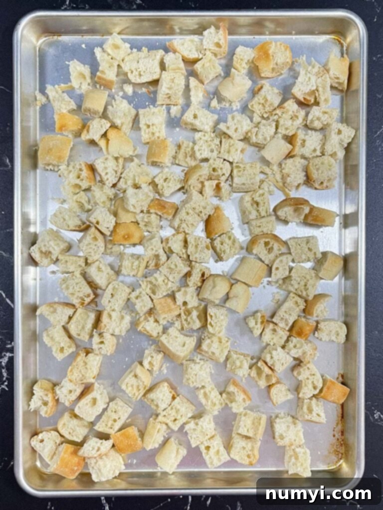 Uniformly cubed ciabatta bread spread on a baking sheet, ready for drying in the oven.